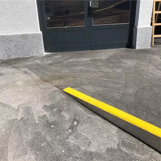 Ein Bereich mit grauem Beton und einer gelben Markierung. Im Hintergrund sind eine Tür und ein Holzregal sichtbar.