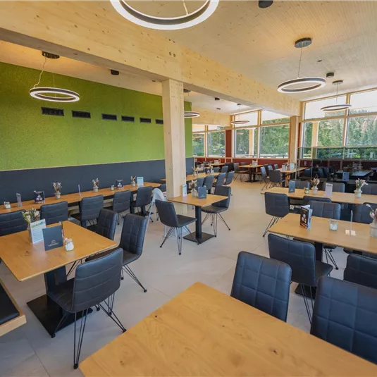 Ein modernes Restaurant mit Holzmöbeln und einem hellen, einladenden Ambiente. Große Fenster sorgen für viel Tageslicht und bieten einen Blick auf die Außenlandschaft.