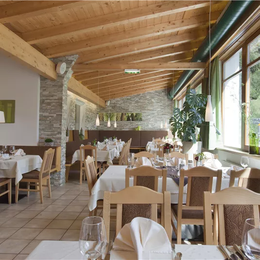 Ein einladendes Restaurant mit Holzbalkendecke und hellen Tischen. Große Fenster bieten einen Blick auf die Natur.