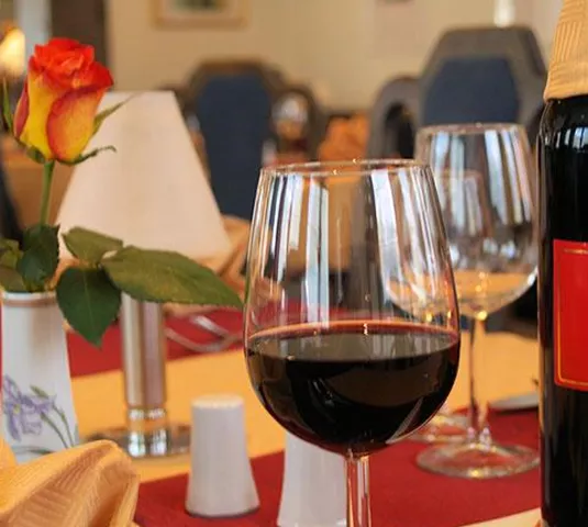 Ein eleganter Tisch mit einem Glas Rotwein, einer Weinflasche und einer Rose in einer Vase. Die Tischdekoration wirkt einladend und gemütlich.