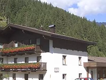 Ein gemütliches Haus mit Balkonen, umgeben von Bergen und Bäumen. Es steht neben einer Straße mit einigen Autos geparkt.