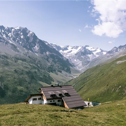 Ein gemütliches Haus liegt in einer grünen Berglandschaft. Majestätische Berge mit Schnee bedeckten Gipfeln umgeben die Szene.