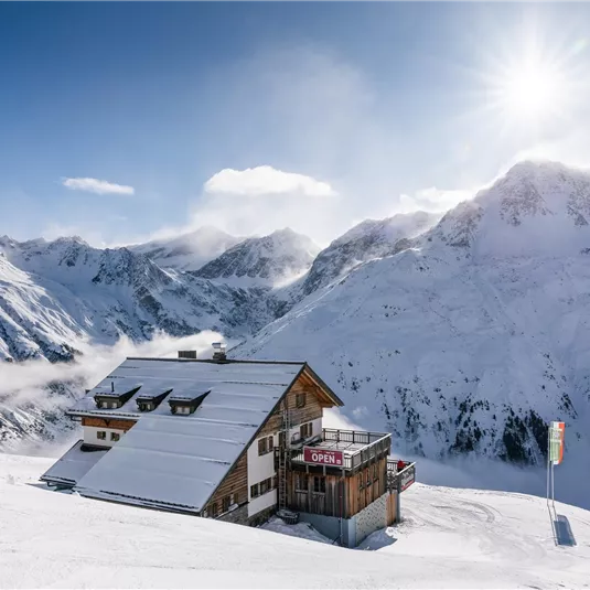 Ein schneebedecktes Bergpanorama mit einer Berghütte im Vordergrund. Die Sonne scheint über den Gipfeln und es herrscht eine klare, kalte Atmosphäre.