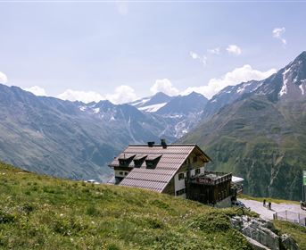 A cabin on a green meadow with impressive mountains in the background. The sky is clear and the landscape appears peaceful and inviting.