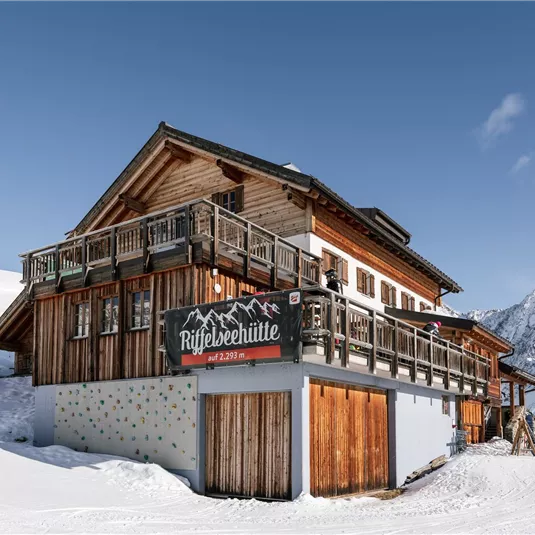 Eine gemütliche Berghütte umgeben von Schnee und Bergen. Der klare Himmel und die sonnige Atmosphäre verleihen dem Ort eine einladende Stimmung.