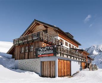 Eine gemütliche Berghütte umgeben von Schnee und Bergen. Der klare Himmel und die sonnige Atmosphäre verleihen dem Ort eine einladende Stimmung.