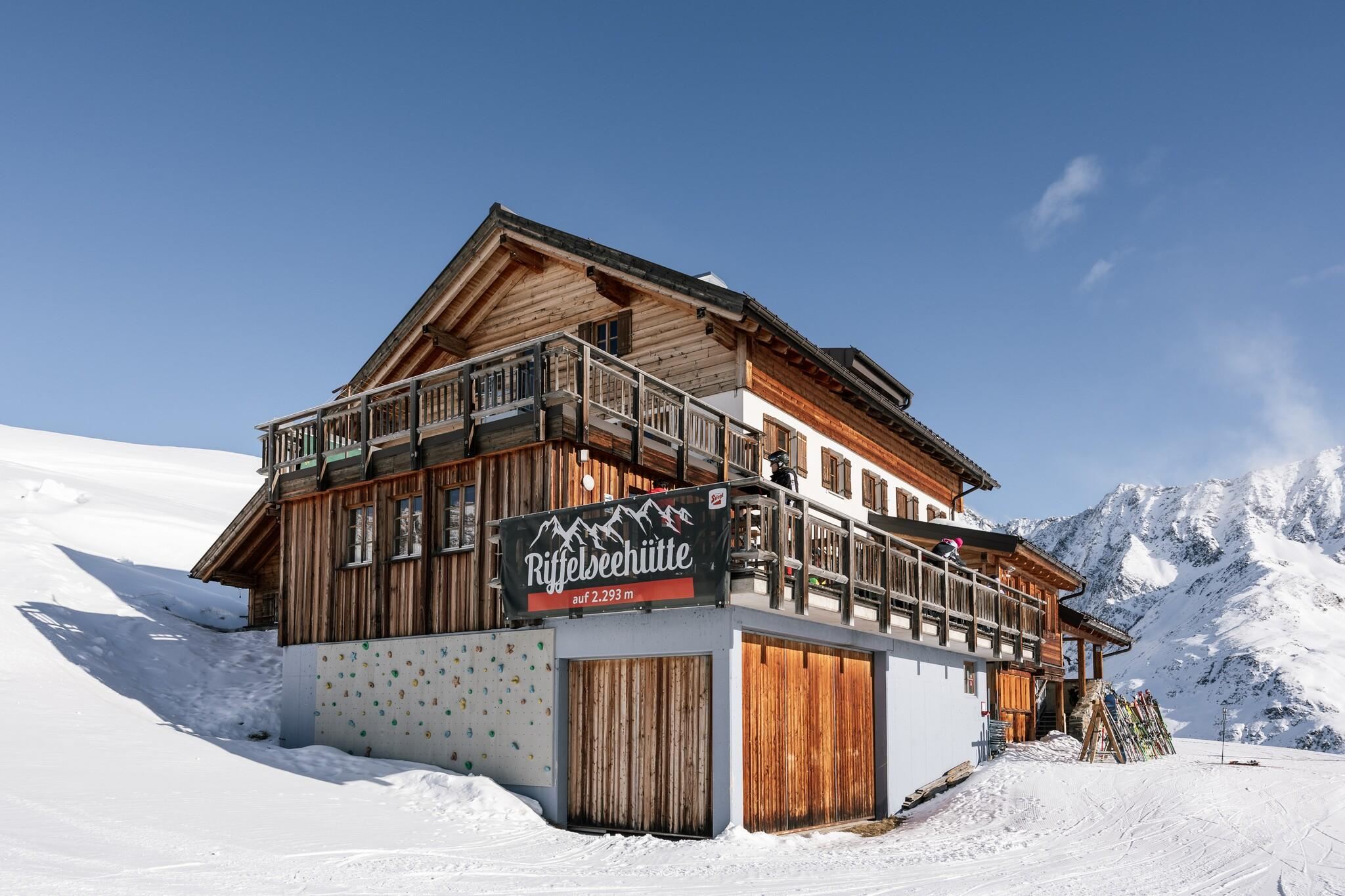 Eine gemütliche Berghütte umgeben von Schnee und Bergen. Der klare Himmel und die sonnige Atmosphäre verleihen dem Ort eine einladende Stimmung.