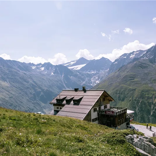A cabin on a green meadow with impressive mountains in the background. The sky is clear and the landscape appears peaceful and inviting.