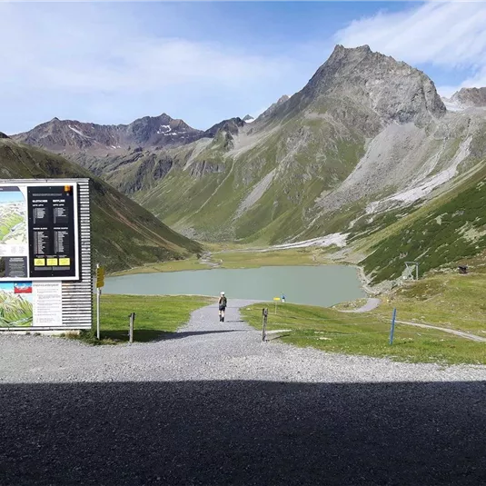 Eine malerische Berglandschaft mit einem klaren See und beeindruckenden Gipfeln im Hintergrund. Ein Wanderer geht entlang eines Weges, während ein Informationsschild auf der linken Seite sichtbar ist.