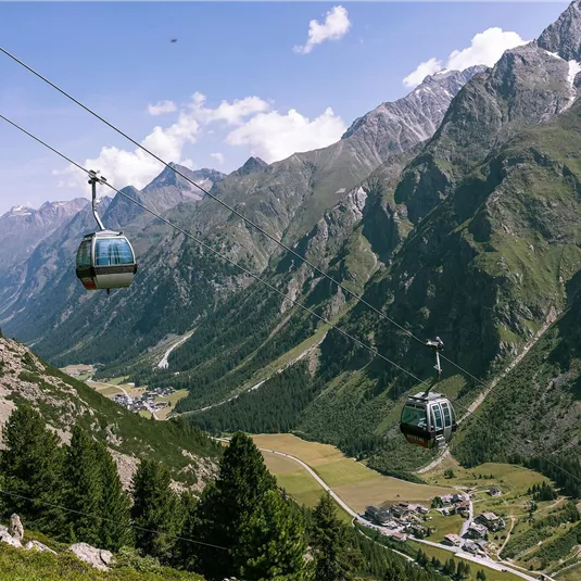 A cable car glides over a picturesque valley with high mountains. In the background, green meadows and a small village can be seen.