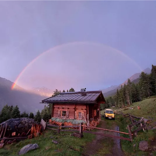 Eine malerische Holzhütte in den Bergen mit einem Regenbogen im Hintergrund. Der Garten ist umgeben von grünen Wiesen und Bäumen.