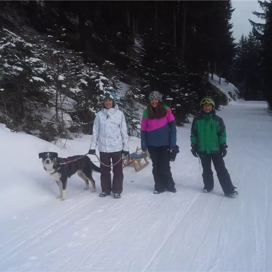 Drei Personen stehen auf einem verschneiten Weg im Wald, begleitet von einem Hund. Die Stimmung ist winterlich, mit Schnee und Tannenbäumen im Hintergrund.