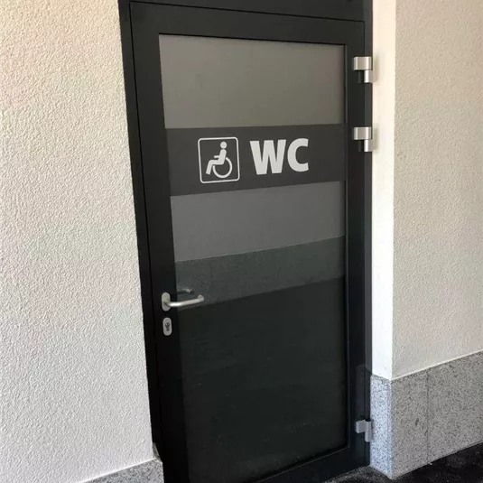 Eine Zugangstür zu einem barrierefreien WC. Die Tür ist modern gestaltet und trägt das Schild "WC" mit einem Symbol für Menschen mit Behinderung.