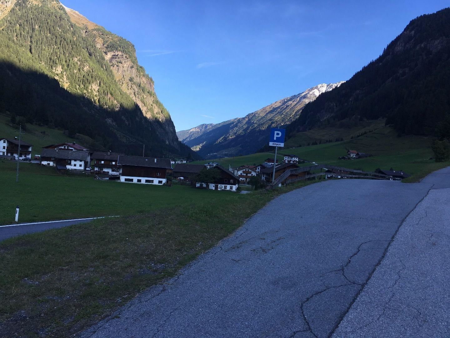 Eine malerische Berglandschaft mit grünen Wiesen und kleinen Häusern im Tal. Im Hintergrund erheben sich hohe Berge unter einem klaren blauen Himmel.