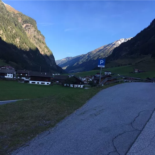 Eine malerische Berglandschaft mit grünen Wiesen und kleinen Häusern im Tal. Im Hintergrund erheben sich hohe Berge unter einem klaren blauen Himmel.