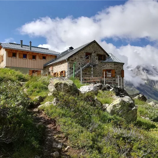 Eine Berghütte umgeben von grünen Wiesen und Felsen. Im Hintergrund sind hohe Berge und Wolken sichtbar.