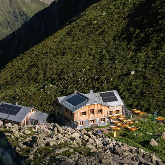 Eine malerische Berghütte umgeben von grünen Hügeln und Felsen. Die Sonne scheint auf die Gebäude mit Solarpanelen.