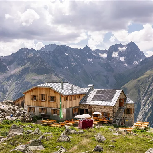 Eine idyllische Berghütte eingebettet in eine beeindruckende Berglandschaft. Die Umgebung ist grün und bewaldet, umgeben von majestätischen Gipfeln.
