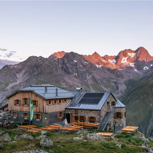 Eine Berghütte inmitten beeindruckender Berge. Die Umgebung ist grün und die Gipfel sind mit Licht erleuchtet.
