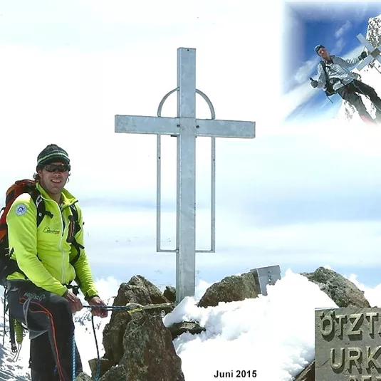 A mountaineer stands at the summit with a cross in the background. The landscape is surrounded by snow-capped mountains.