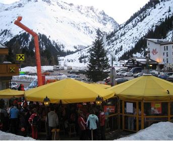 Ein gemütliches Restaurant mit gelben Sonnenschirmen in einer schneebedeckten Berglandschaft. Im Hintergrund sind schneebedeckte Berge und ein Parkplatz zu sehen.