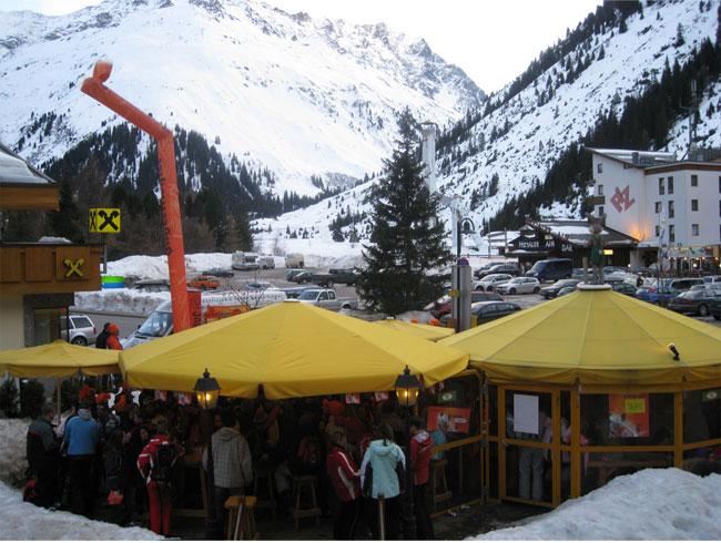 Ein gemütliches Restaurant mit gelben Sonnenschirmen in einer schneebedeckten Berglandschaft. Im Hintergrund sind schneebedeckte Berge und ein Parkplatz zu sehen.