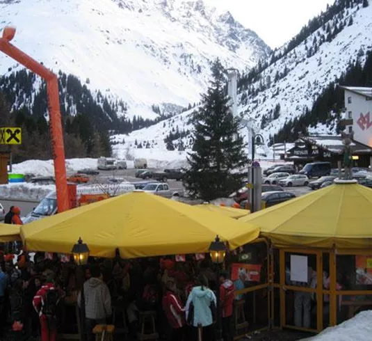 Ein gemütliches Restaurant mit gelben Sonnenschirmen in einer schneebedeckten Berglandschaft. Im Hintergrund sind schneebedeckte Berge und ein Parkplatz zu sehen.