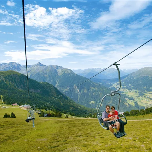 Eine Seilbahnfahrt durch die Berge mit grünen Wiesen und blauer Himmel. Zwei Personen genießen die Aussicht auf die Landschaft.