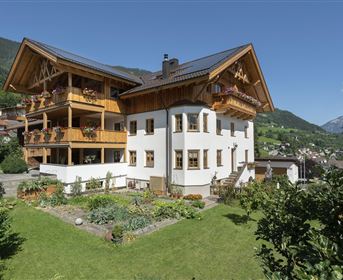 Ein schönes, modernes Gebäude im alpenländischen Stil mit Balkonen und großen Fenstern. Der Garten ist gepflegt und die Umgebung ist von Bergen umgeben.