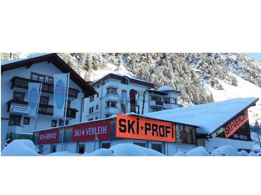 Ein Ski-Verleih mit dem Schild „SKI-PROFI“ in einer schneebedeckten Umgebung. Im Hintergrund sind Berglandschaften und ein traditionelles Gebäude zu sehen.