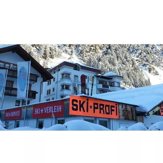 Ein Ski-Verleih mit dem Schild „SKI-PROFI“ in einer schneebedeckten Umgebung. Im Hintergrund sind Berglandschaften und ein traditionelles Gebäude zu sehen.