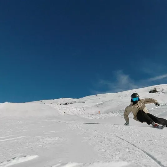 Ein Skifahrer fährt mit Schwung über die Schneepiste. Der Himmel ist klar und blau, und die Landschaft ist von weißem Schnee bedeckt.