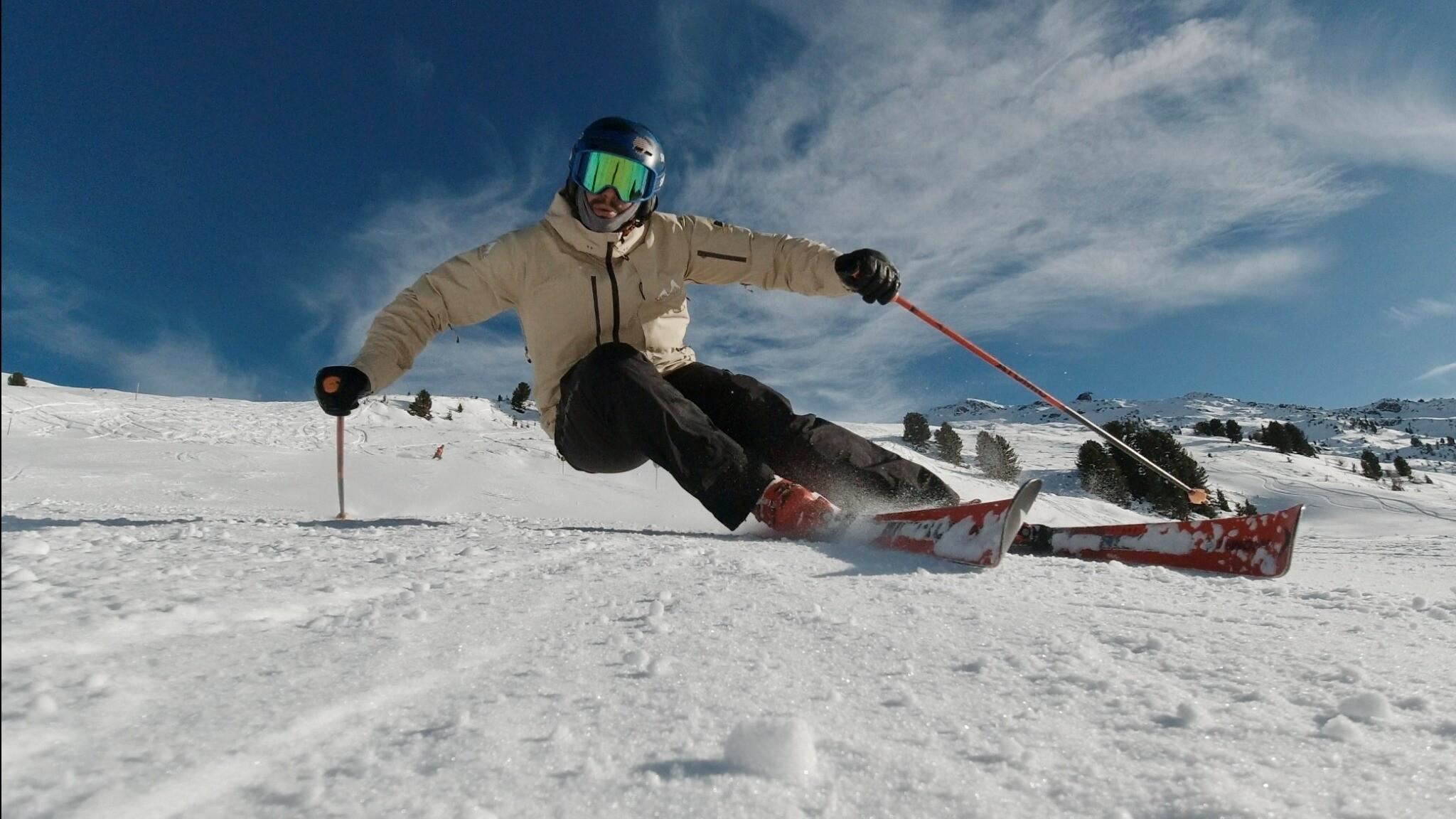 Ein Skifahrer fährt über frischen Schnee auf einer sonnigen Piste. Im Hintergrund sind schneebedeckte Berge und ein blauer Himmel sichtbar.