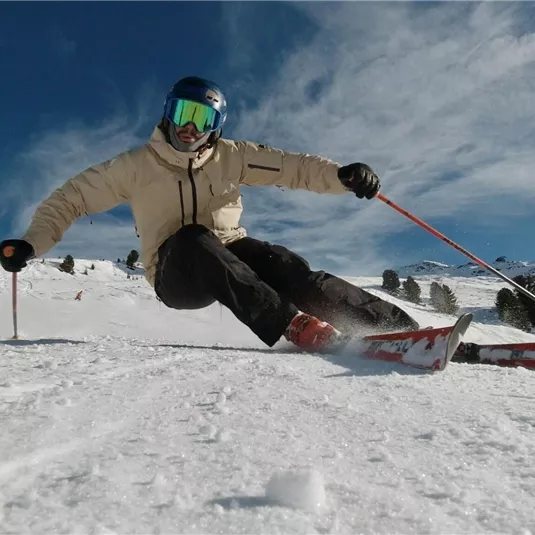Ein Skifahrer fährt über frischen Schnee auf einer sonnigen Piste. Im Hintergrund sind schneebedeckte Berge und ein blauer Himmel sichtbar.