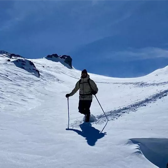 Eine Person wandert durch eine verschneite Landschaft mit Skistock. Im Hintergrund sind andere Wanderer auf einem Berg sichtbar.