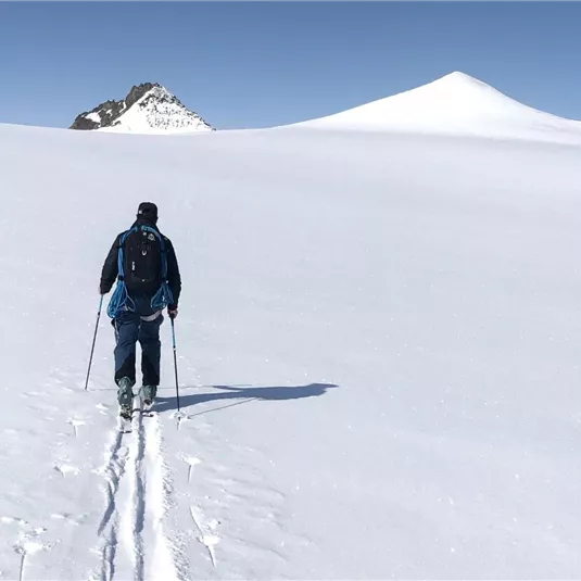 Ein einzelner Wanderer geht durch eine weite, schneebedeckte Landschaft. Im Hintergrund ist ein schneebedeckter Gipfel unter klarem Himmel zu sehen.