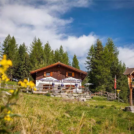 Eine gemütliche Berghütte umgeben von grünen Wiesen und Bäumen. Der Himmel ist blau mit ein paar Wolken.