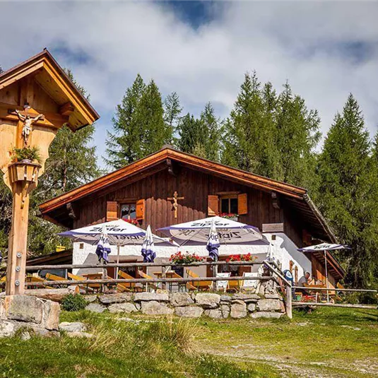 Eine gemütliche Berghütte umgeben von Tannenbäumen und Wiesen. Auf der Terrasse stehen Tische mit Sonnenschirmen, die zum Verweilen einladen.