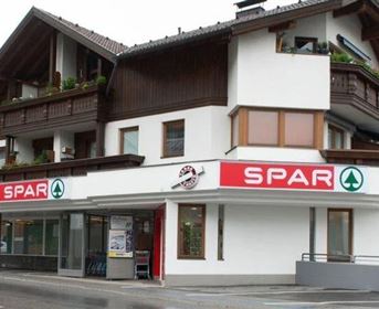 Ein modernes Geschäft mit einem SPAR-Schild im Erdgeschoss. Das Gebäude hat einen traditionellen alpinen Stil mit Bal­kons und bunten Pflanzen.