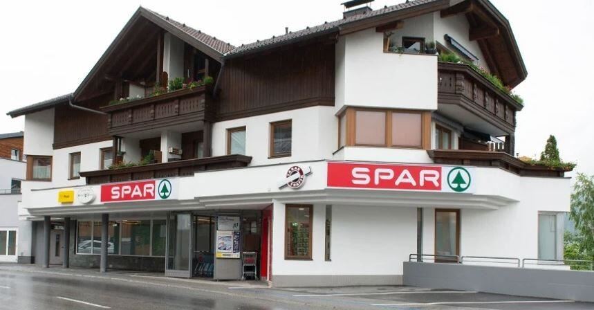 Ein modernes Gebäude mit einem SPAR-Supermarkt im Erdgeschoss. Die Fassade hat einen alpenländischen Stil mit Balkonen und Pflanzen.