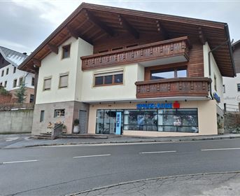 A modern building with wooden cladding and a large window on the ground floor. It is located on a street and has a clear sky in the background.