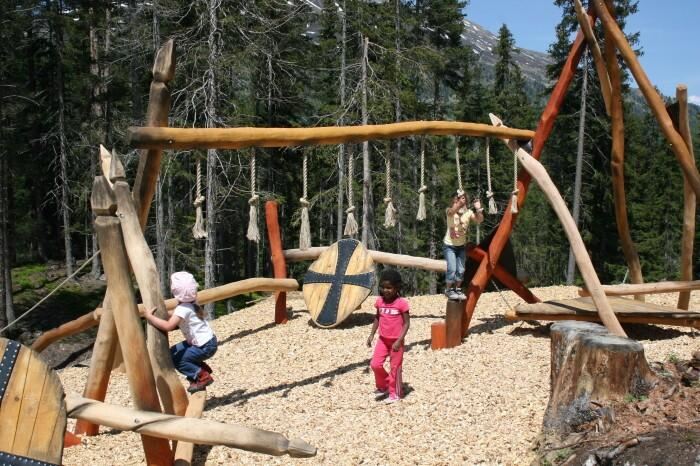 A playground in the forest with wooden play equipment. Children are joyfully playing and climbing in nature.