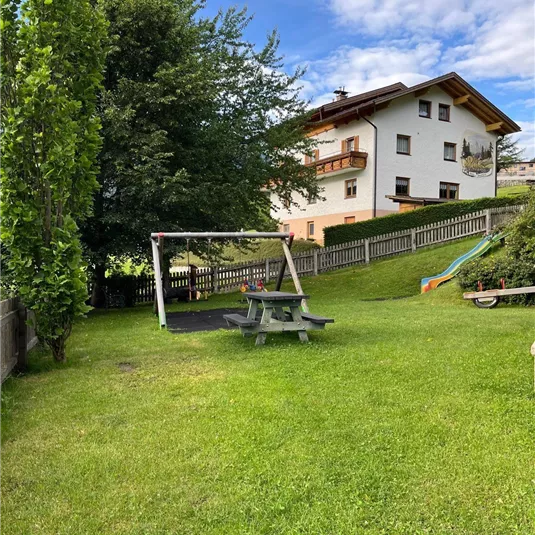 Ein gepflegter Garten mit einer Schaukel und einem Picknicktisch. Im Hintergrund steht ein schönes Haus und der Himmel ist teilweise bewölkt.