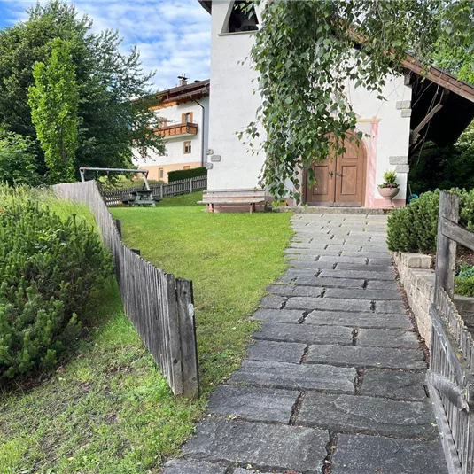 Ein schöner Weg aus Stein führt zu einem charmanten Gebäude mit weißer Fassade. Umgeben von grünen Bäumen und Sträuchern, strahlt die Szene eine ruhige und einladende Atmosphäre aus.