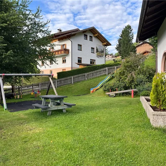 Ein gepflegter Garten mit einer Schaukel und einer Rutsche. Im Hintergrund sind ein paar Wohnhäuser und Bäume zu sehen.