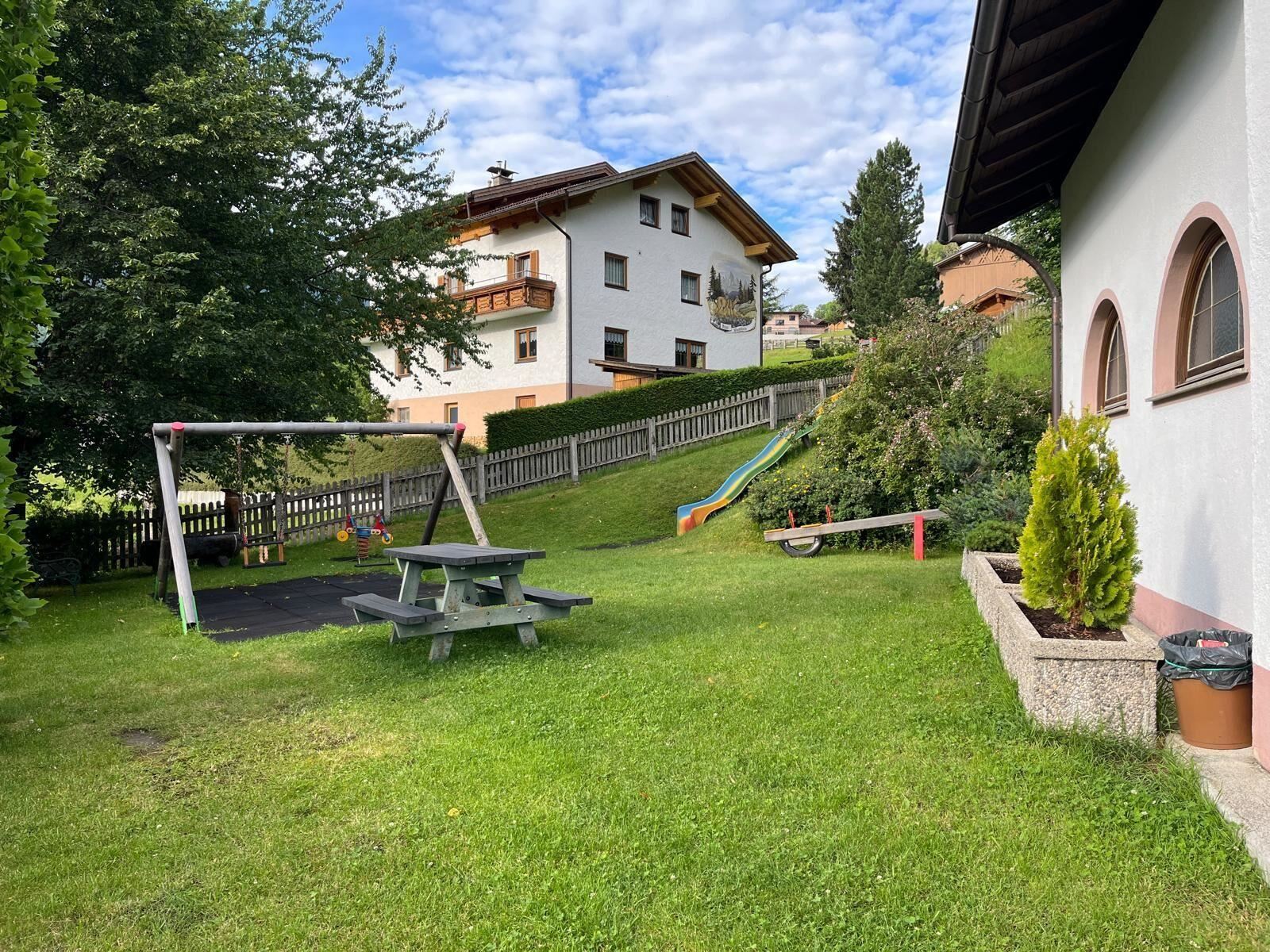 Ein gepflegter Garten mit einer Schaukel und einer Rutsche. Im Hintergrund sind ein paar Wohnhäuser und Bäume zu sehen.