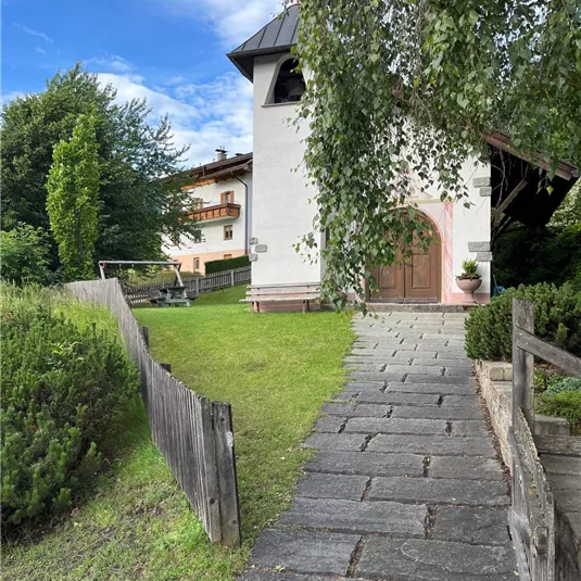 Ein schöner Weg führt zur Kirche, umgeben von üppigem Grün. Im Hintergrund sind weitere Gebäude in einer ruhigen Umgebung zu sehen.