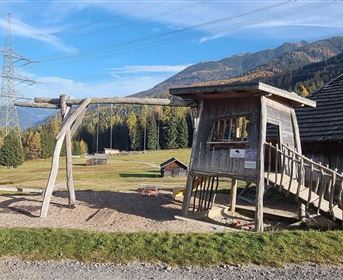 A playground with a wooden swing and a playhouse in a rural area. In the background, there are trees and mountains under a clear blue sky.