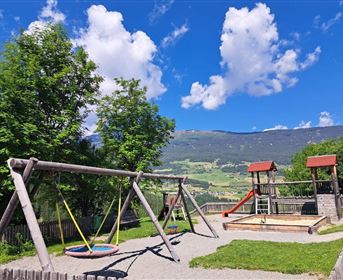 A playground with a swing and slides, surrounded by trees and lush meadows. The sky is clear and sunny with a few clouds.