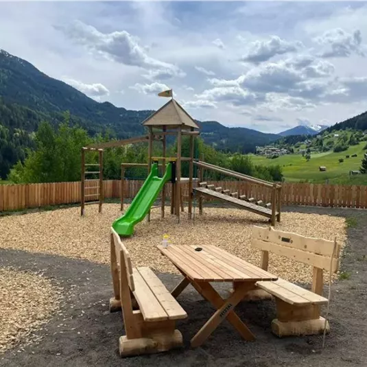 Ein Spielplatz mit einer Rutsche und Klettergerüst in einer naturnahen Umgebung. Im Hintergrund sind Berge und ein blauer Himmel zu sehen.
